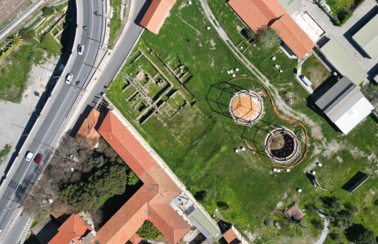 Foto aerea di un'area archeologica con resti di edifici romani e di due gasometri metallici. Il terreno è un grande prato verde intenso. Sulla sinistra passa una strada con auto e oltre la strada si vede la prosecuzione dell'area archeologica. Ai margini si vedono i tetti in tegole di costruzioni moderne.